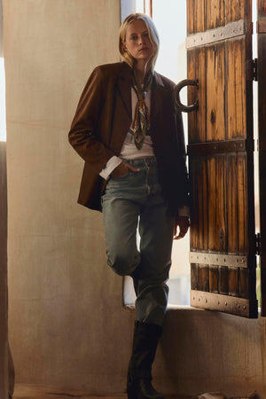 Wearing the SALIE FAUX SUEDE JACKET by Velvet by Graham & Spencer over a white shirt and patterned scarf with blue jeans and black boots, this person stands by an open wooden door in a softly lit room—a timeless layering piece for any occasion.