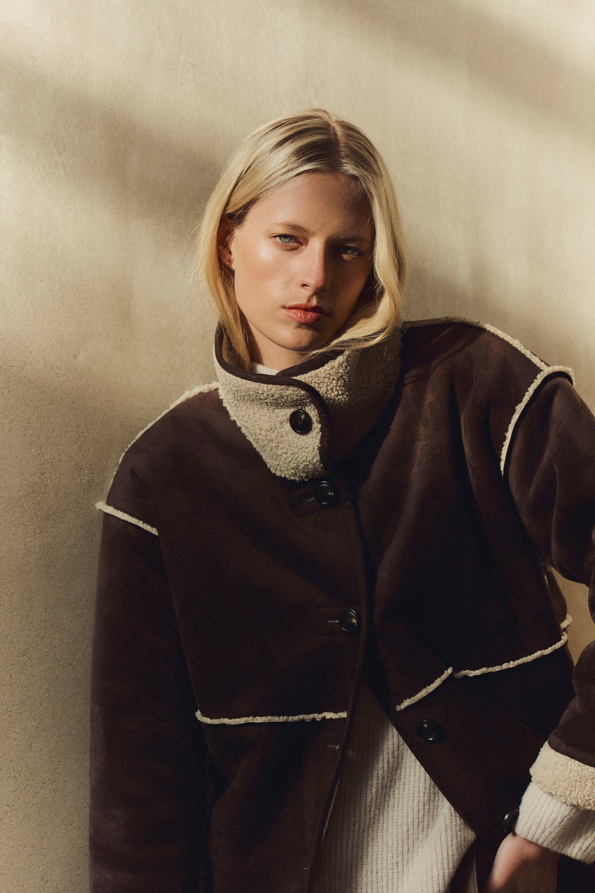 A woman with straight blonde hair wears the SHONA SHERPA REVERSIBLE JACKET by Velvet by Graham & Spencer over a cream sweater, standing against a beige textured wall in soft lighting.-39814925877441