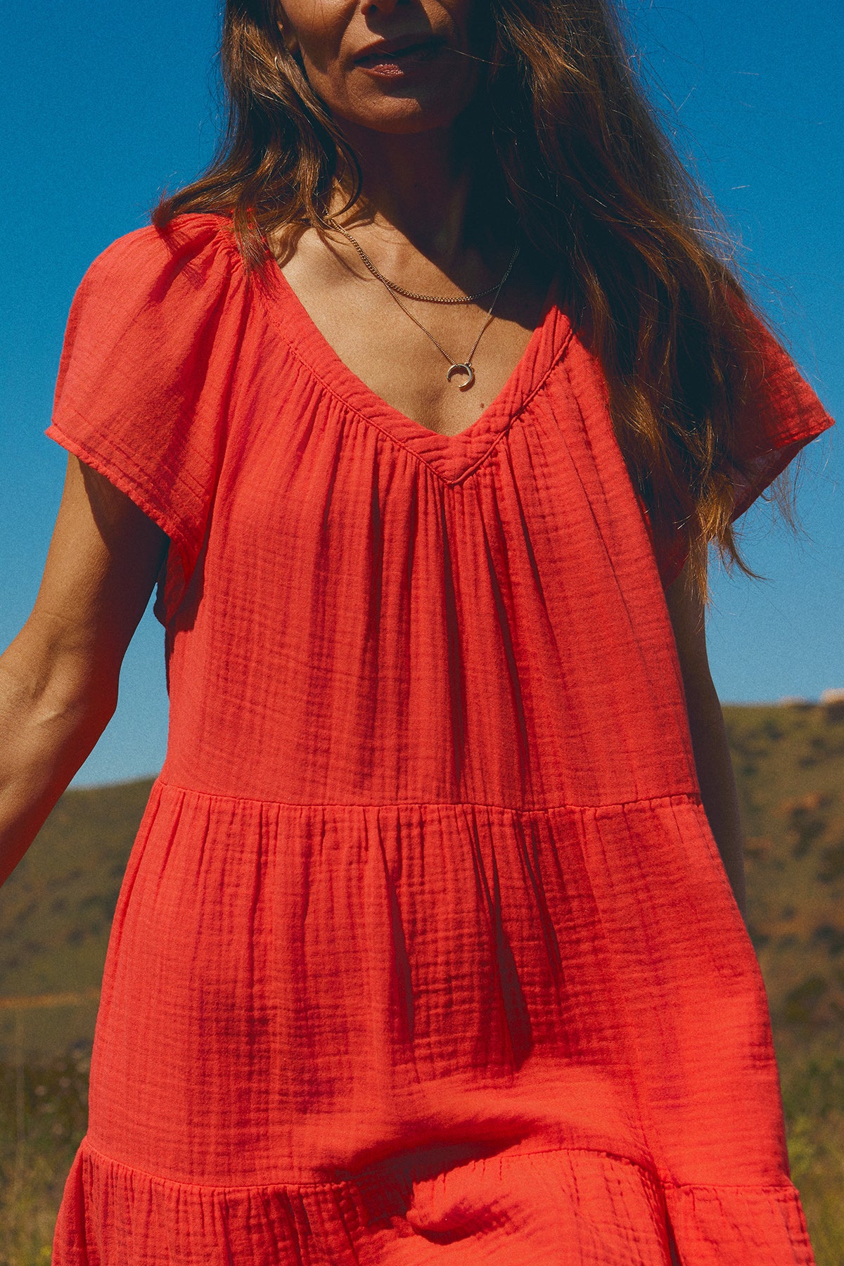A woman in the Eleanor Cotton Gauze Dress by Velvet by Graham & Spencer stands outdoors against a backdrop of blue sky. The lower half of her face is visible. The airy cotton gauze fabric and tiered silhouette add a touch of effortless elegance to the scene.