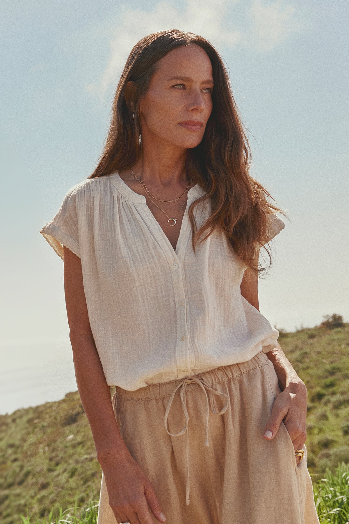 A woman with long brown hair wearing the PAMELA COTTON GAUZE BUTTON-UP TOP from Velvet by Graham & Spencer and beige drawstring pants stands outdoors on a grassy hill, enjoying a spring weekend.-37618811011265