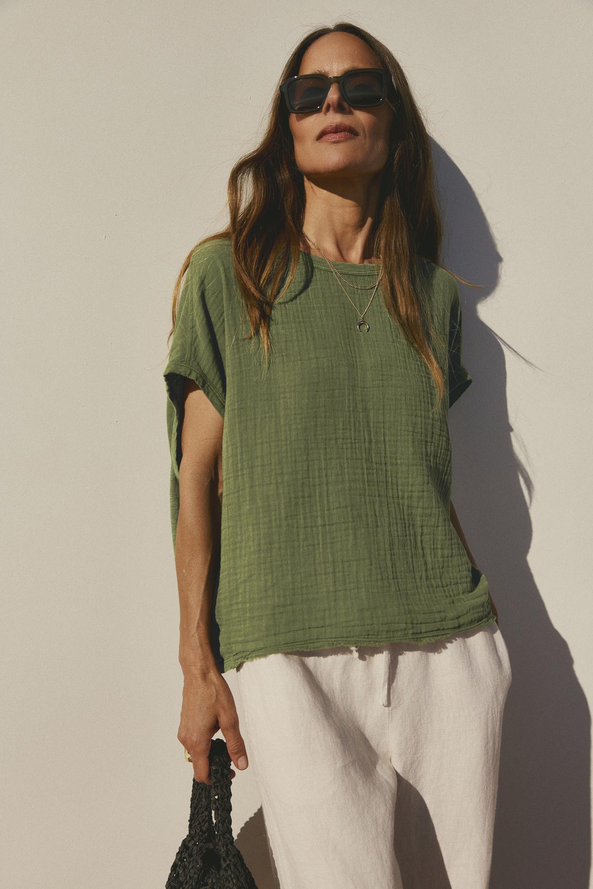 Woman in a green Velvet by Graham & Spencer Caroline Cocoon Top and white pants standing against a white wall, wearing sunglasses and holding a black bag.-36533032714433