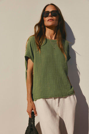 Woman in a green Velvet by Graham & Spencer Caroline Cocoon Top and white pants standing against a white wall, wearing sunglasses and holding a black bag.