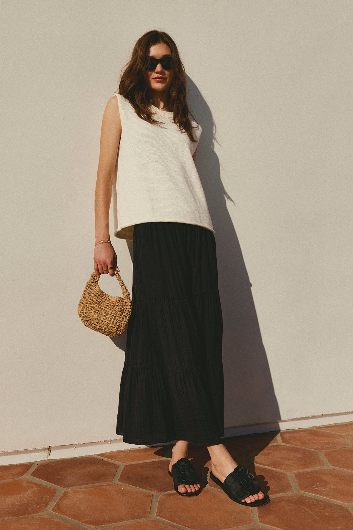   A woman in sunglasses, a white sleeveless top, and a DANIELLE COTTON GAUZE TIERED SKIRT from Velvet by Graham & Spencer stands on a tiled surface, holding a straw bag. 