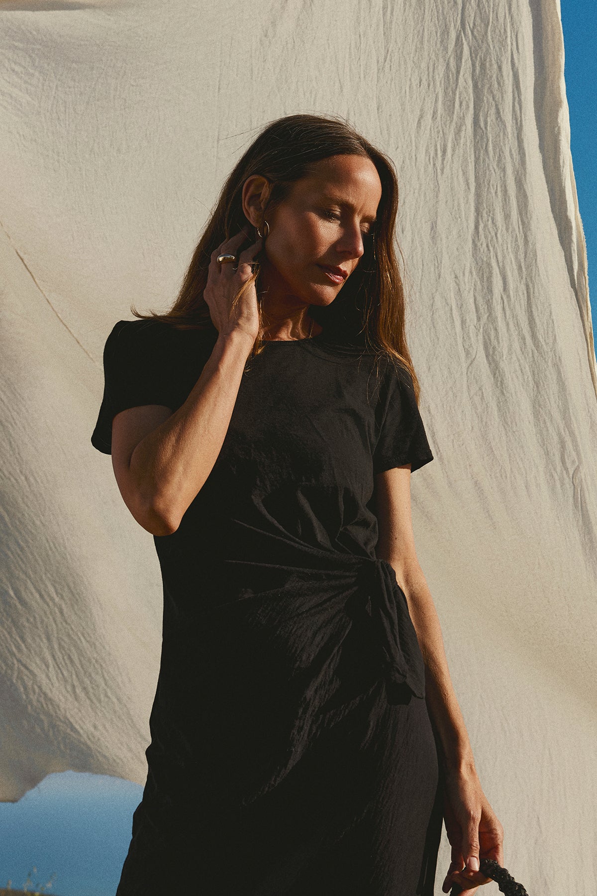 Woman in black DARCY COTTON SLUB MIDI DRESS by Velvet by Graham & Spencer posing thoughtfully with hand on her earring, standing against a draped white cloth backdrop.-36783604105409