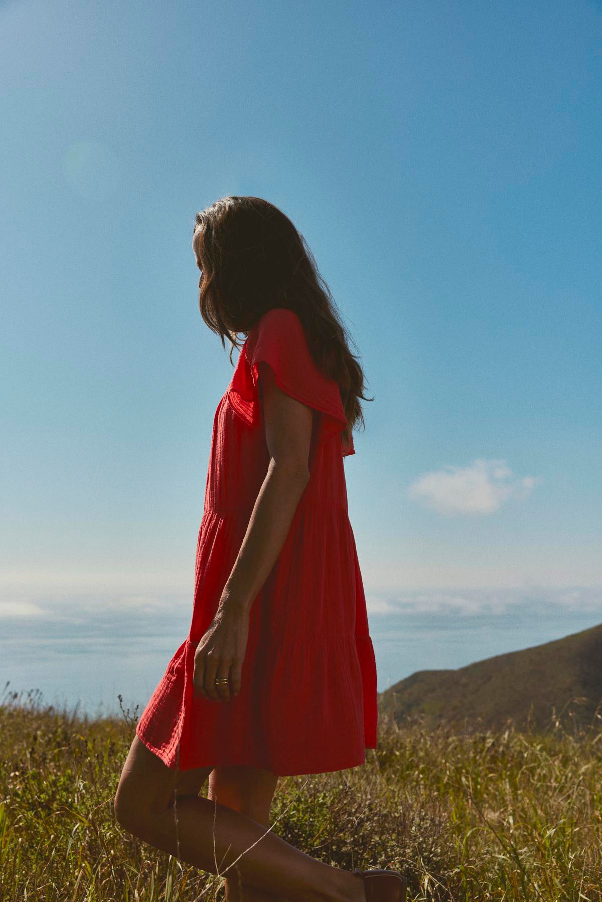 A woman in an ELEANOR COTTON GAUZE TIERED DRESS by Velvet by Graham & Spencer walking through a field with a clear blue sky and hills in the background.-36532828700865