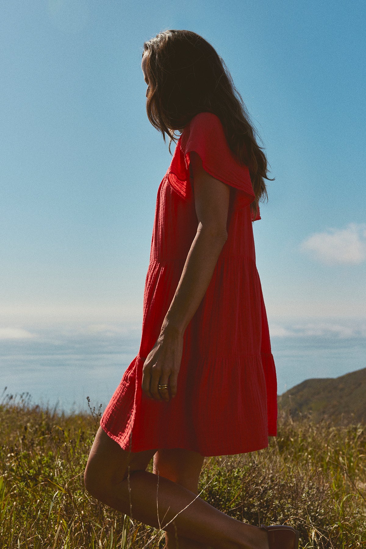 A person wearing the ELEANOR COTTON GAUZE DRESS by Velvet by Graham & Spencer stands in a grassy field overlooking a distant body of water under a clear blue sky.-37618633965761