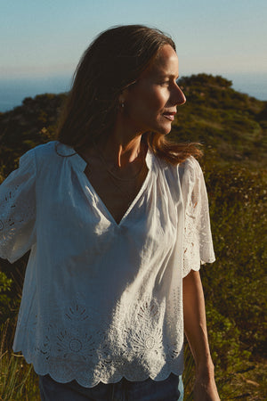 Woman in a white Velvet by Graham & Spencer RAZI EMBROIDERED COTTON LACE TOP with scallop cuffs and hem, looking to the side in a sunny, natural setting with shrubs and the sea in the background.