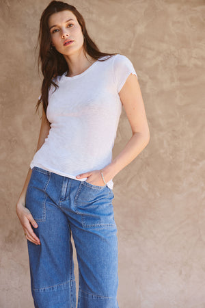 A woman stands against a beige textured wall, wearing the Velvet by Graham & Spencer ODELIA TEE in white cotton slub fabric and blue jeans, one hand in her pocket as she looks at the camera.