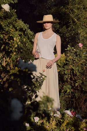 In a garden, a person in a wide-brim hat and beige skirt wears the AYA TANK TOP by Velvet by Graham & Spencer, gently holding a small flower.