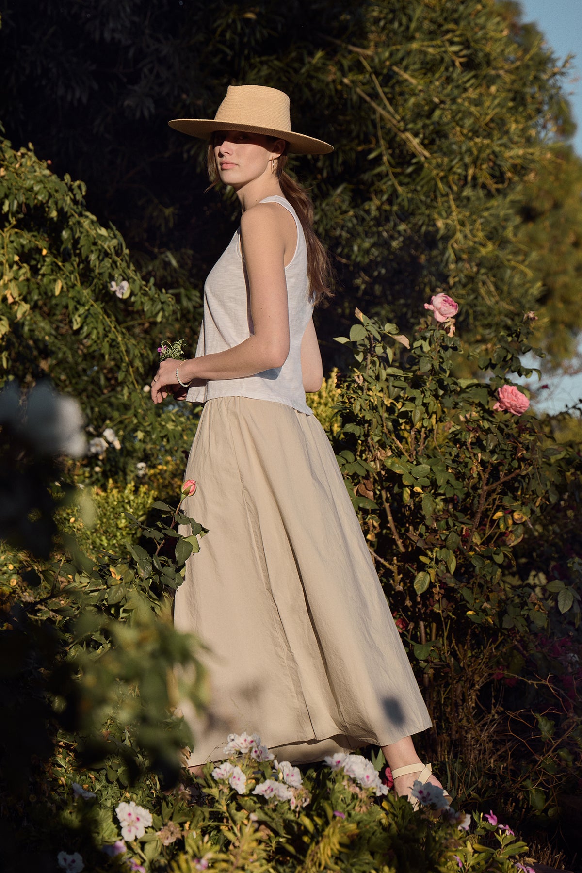 A woman wearing a wide-brimmed hat and the AYA Tank Top by Velvet by Graham & Spencer, expertly layered over her beige outfit, strolls through a garden blooming with lush green foliage and pink flowers under the sunny sky.-38957693075649