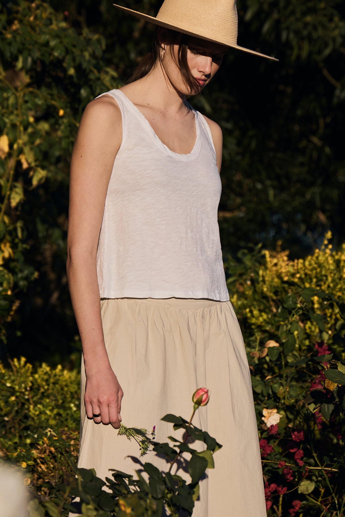 A woman in a garden wears the AYA TANK TOP by Velvet by Graham & Spencer—a white sleeveless top with a scoop neckline—and a beige skirt, accessorized with a wide-brimmed hat. She stands holding a small flower, her effortless style enhancing the serene setting.