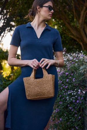 A woman in the FABIANA DRESS by Velvet by Graham & Spencer, a versatile blue outfit, stands outdoors holding a woven handbag next to flowering bushes.