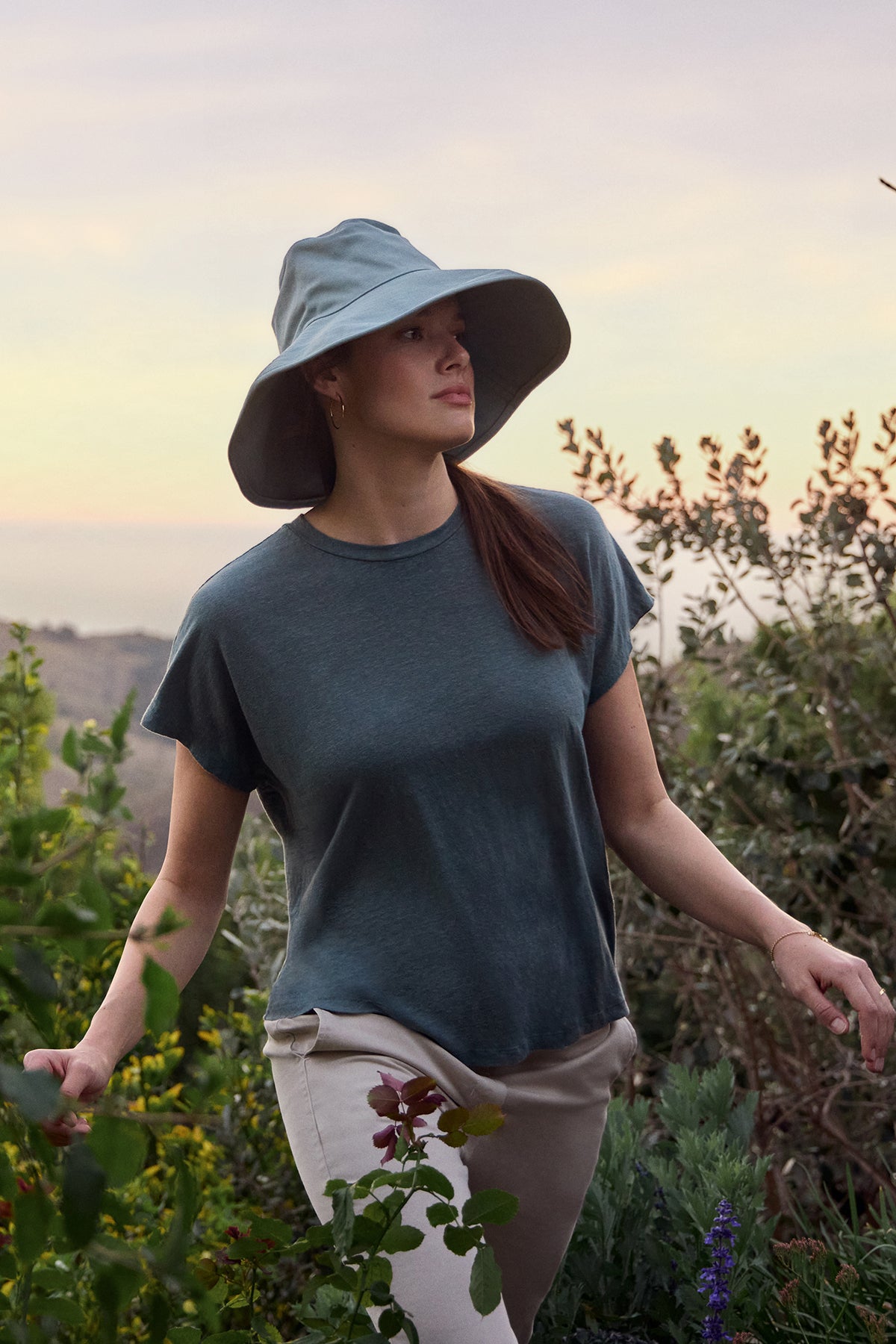 Wearing the JASMINE LINEN KNIT TEE by Velvet by Graham & Spencer, a person strolls through a lush garden under a gentle sky, showcasing the charm of this versatile staple.