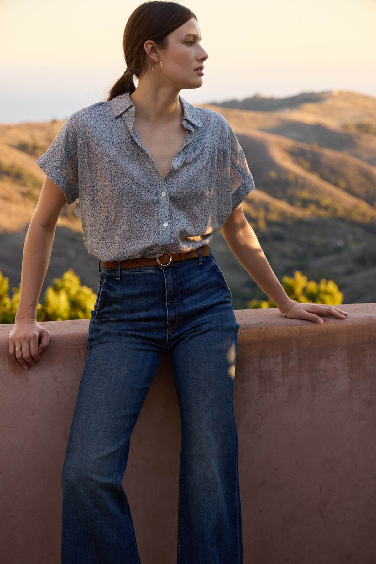 Dressed in a Velvet by Graham & Spencer TIGE printed crinkle cotton gauze top and jeans, a person leans against a ledge, gazing into the distance. The hilly landscape and soft evening light offer a calming backdrop.
