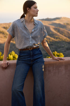 Dressed in a Velvet by Graham & Spencer TIGE printed crinkle cotton gauze top and jeans, a person leans against a ledge, gazing into the distance. The hilly landscape and soft evening light offer a calming backdrop.