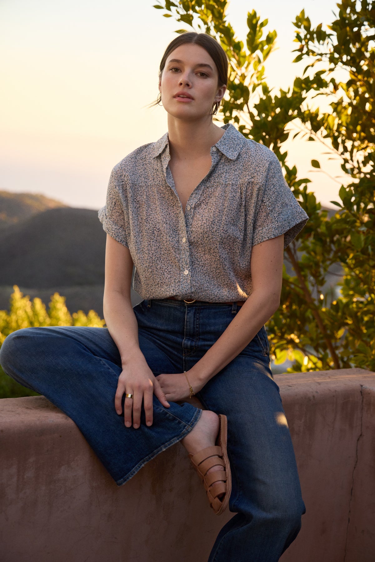 A person sits casually on a stone ledge outdoors, wearing the TIGE Printed Crinkle Cotton Gauze Top by Velvet by Graham & Spencer, blue jeans, and sandals. Greenery and hills are visible in the background during sunset.