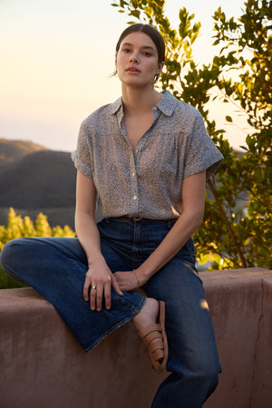 A person sits casually on a stone ledge outdoors, wearing the TIGE Printed Crinkle Cotton Gauze Top by Velvet by Graham & Spencer, blue jeans, and sandals. Greenery and hills are visible in the background during sunset.