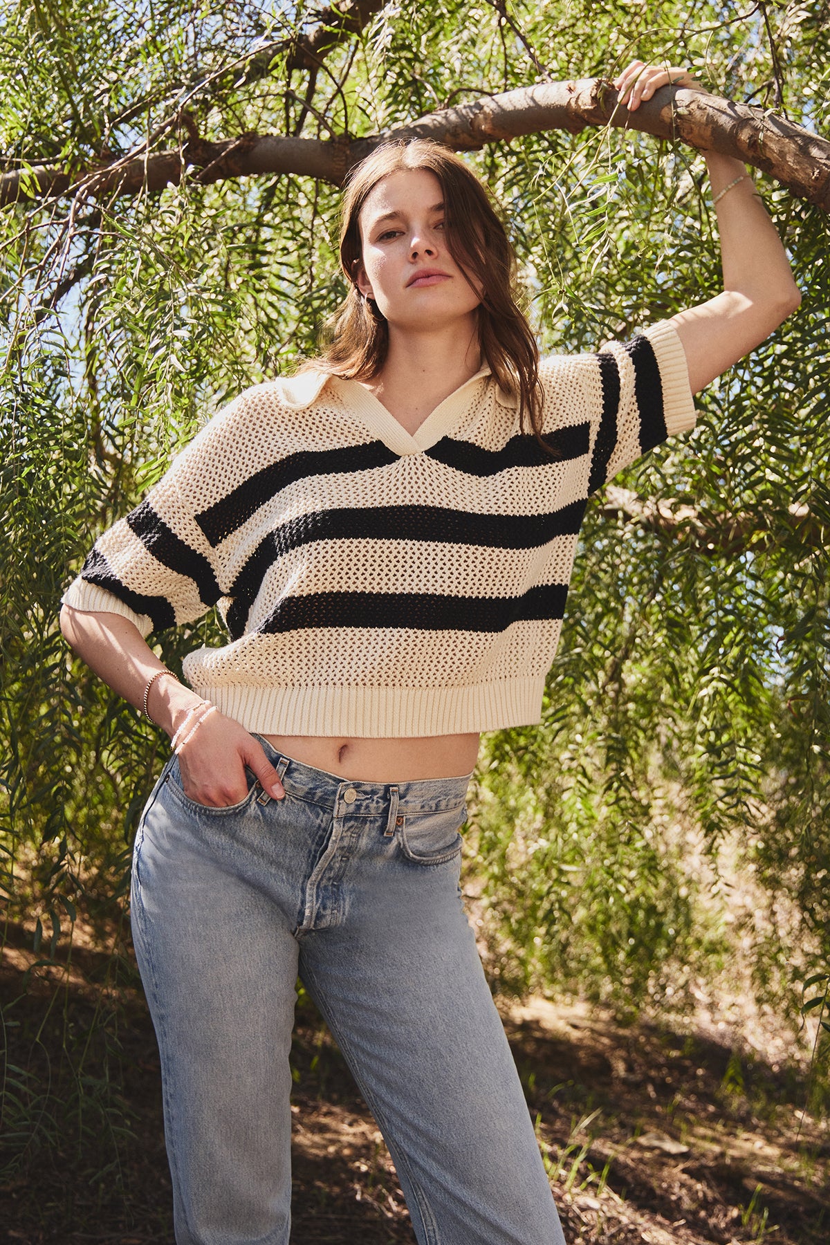 A person in a comfortable YORK COTTON CASHMERE MESH SWEATER POLO by Velvet by Jenny Graham and jeans stands under a tree, with one arm resting on a branch, set against lush greenery.