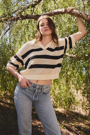 A person in a comfortable YORK COTTON CASHMERE MESH SWEATER POLO by Velvet by Jenny Graham and jeans stands under a tree, with one arm resting on a branch, set against lush greenery.