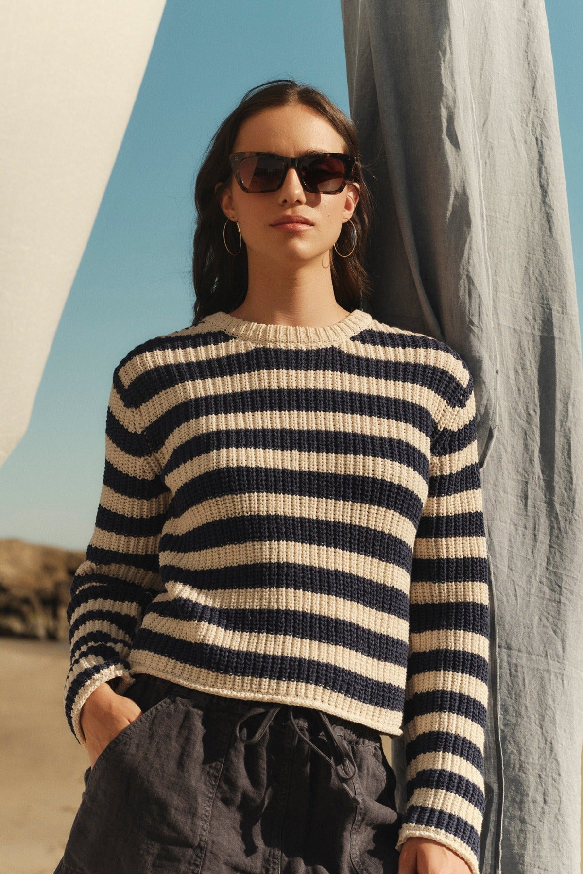A woman wearing a MAXINE SWEATER from Velvet by Graham & Spencer and sunglasses stands confidently in front of a draped fabric background under a clear sky.-36752771121345