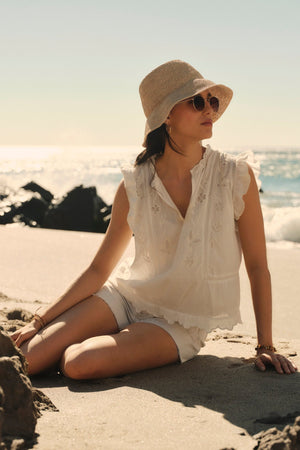 Woman in a white CHARLENE TOP by Velvet by Graham & Spencer embroidery outfit and sunhat sitting on a beach with rocks, looking away from the camera, ocean in the background.