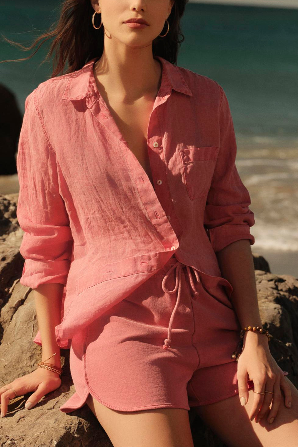 A woman in a pink, Velvet by Graham & Spencer NATALIA LINEN BUTTON-UP SHIRT outfit sitting on a rocky beach shore, looking at the camera with the sea and clear sky in the background.