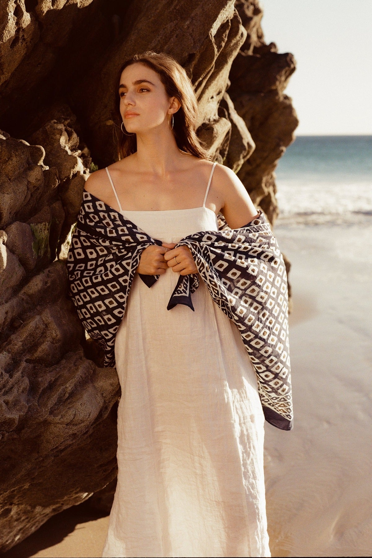   A woman in a Velvet by Graham & Spencer STEPHIE LINEN DRESS and draped patterned shawl stands by a rocky beach, gazing thoughtfully away from the camera. 