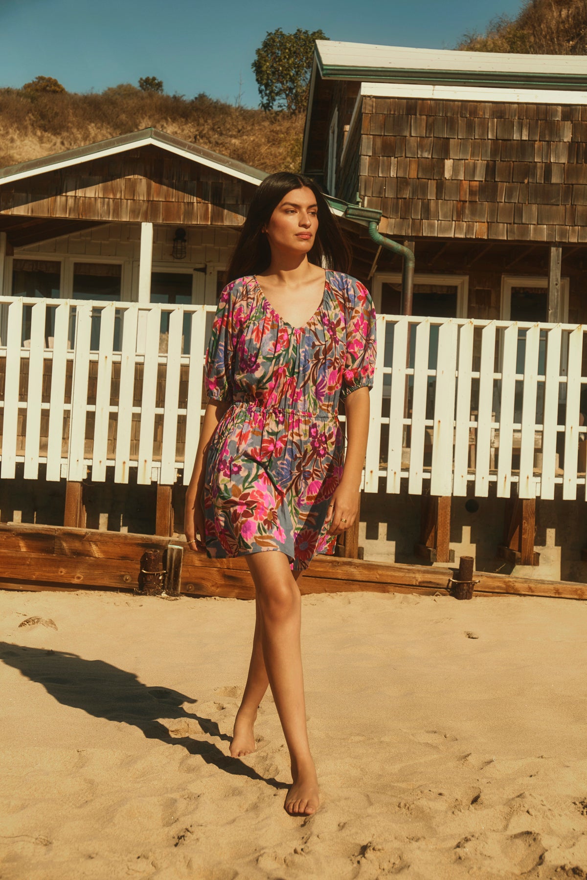 A woman in the HAZEL DRESS by Velvet by Graham & Spencer, a colorful floral style with an elastic waist, walks barefoot on sand near a wooden building with a white fence.-39111856652481