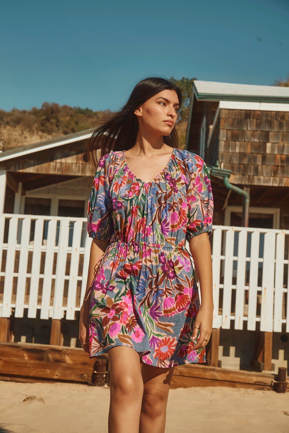 A woman walks outdoors on a sunny day, wearing the HAZEL DRESS by Velvet by Graham & Spencer—a colorful floral dress with puff sleeves and an elastic waist—against a backdrop of wooden buildings and a white fence.-39111856521409