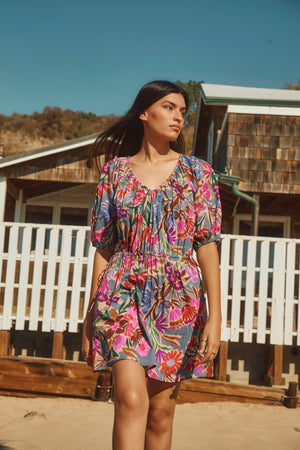 A woman walks outdoors on a sunny day, wearing the HAZEL DRESS by Velvet by Graham & Spencer—a colorful floral dress with puff sleeves and an elastic waist—against a backdrop of wooden buildings and a white fence.