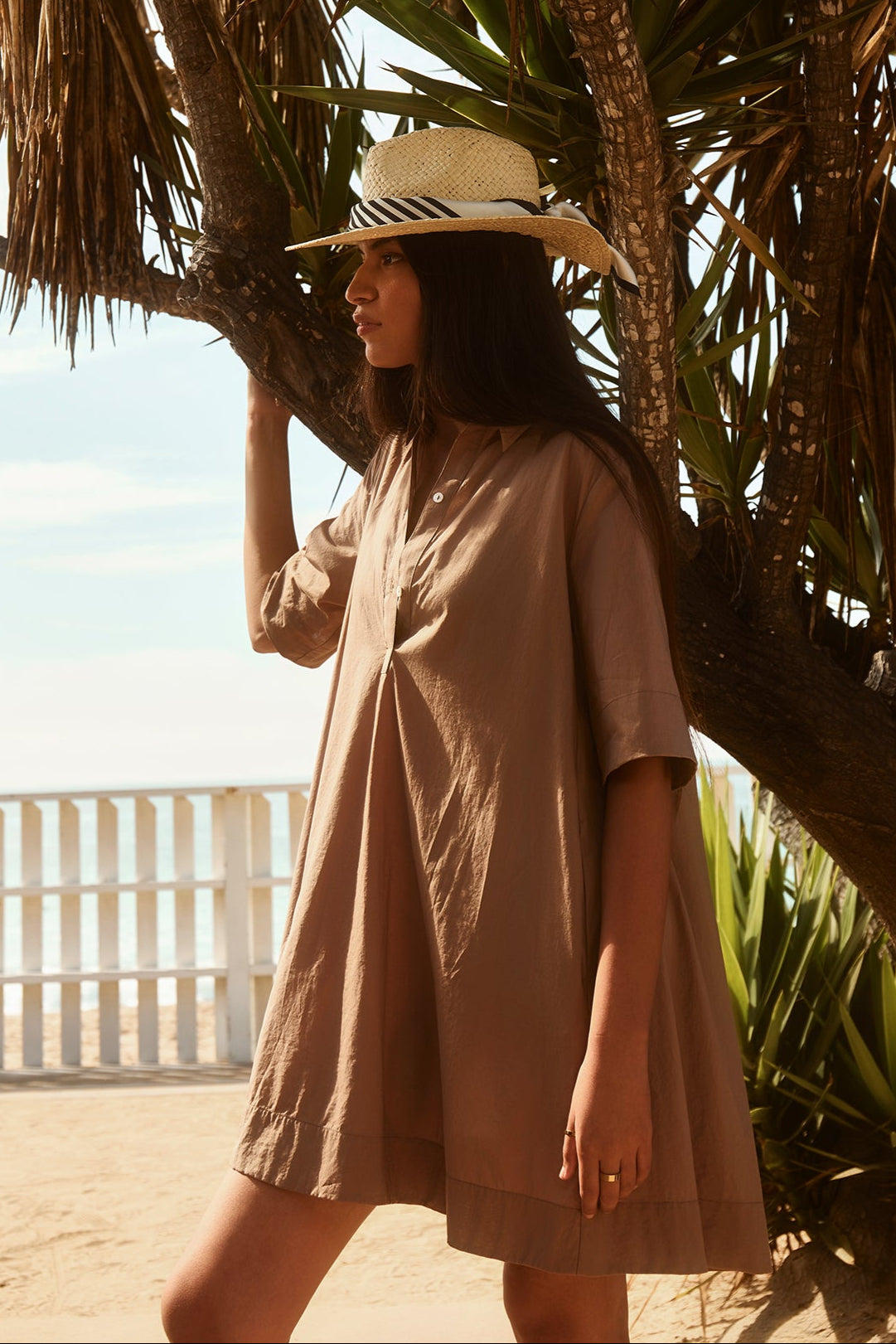 A woman in the DRIANA DRESS by Velvet by Graham & Spencer, a loose brown cotton poplin style with side seam pockets, paired with a straw hat, stands under a tree near a white fence at the beach.