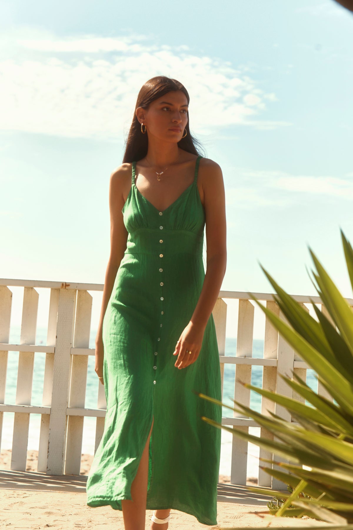 A woman wears the Velvet by Graham & Spencer JANET LINEN DRESS, a sleeveless green woven linen tank dress, while standing near a white wooden fence on a sunny beach with the ocean and sky behind her.-39119904506049