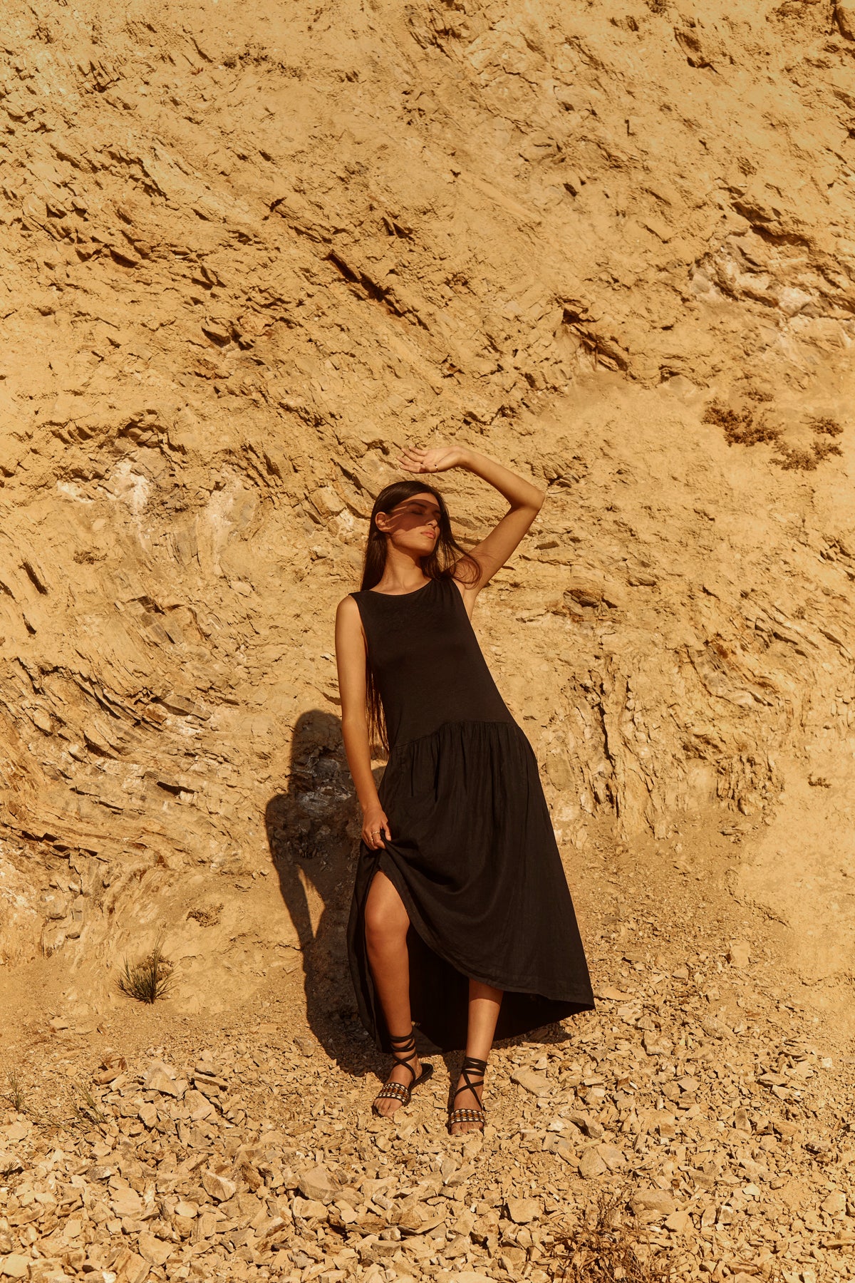 Wearing the MABLE LINEN KNIT DRESS by Velvet by Graham & Spencer, a woman stands on rocky ground before a beige cliff, shielding her eyes from the sun with her hand.