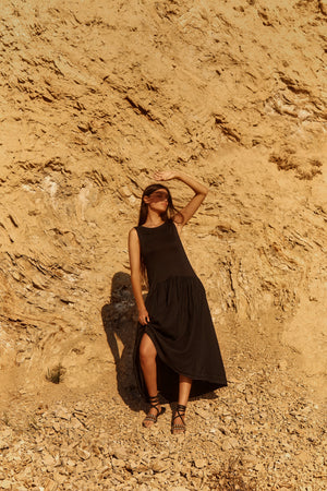 Wearing the MABLE LINEN KNIT DRESS by Velvet by Graham & Spencer, a woman stands on rocky ground before a beige cliff, shielding her eyes from the sun with her hand.