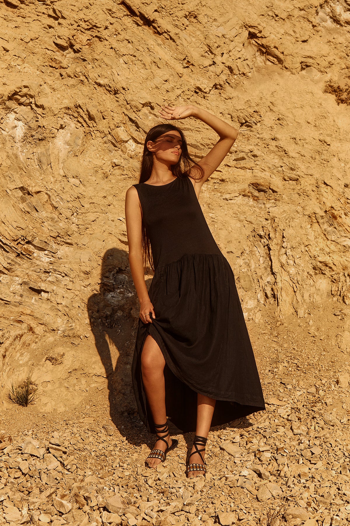 A woman in the MABLE LINEN KNIT DRESS by Velvet by Graham & Spencer pairs it with sandals as she stands on rocky terrain, shading her eyes from the sun with her hand.-39320056987841