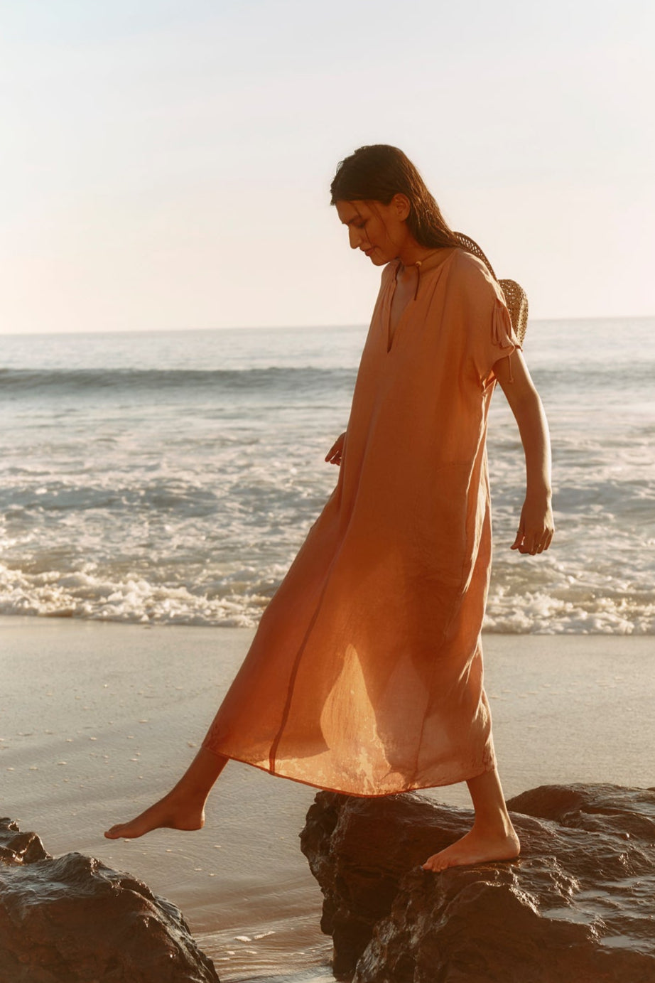 Wearing the MAEVE LINEN DRESS by Velvet by Graham & Spencer, a person strolls barefoot on seaside rocks as gentle waves roll in—a perfect scene of effortless summer style.