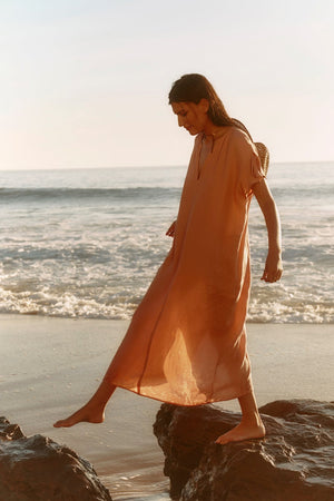 Wearing the MAEVE LINEN DRESS by Velvet by Graham & Spencer, a person strolls barefoot on seaside rocks as gentle waves roll in—a perfect scene of effortless summer style.