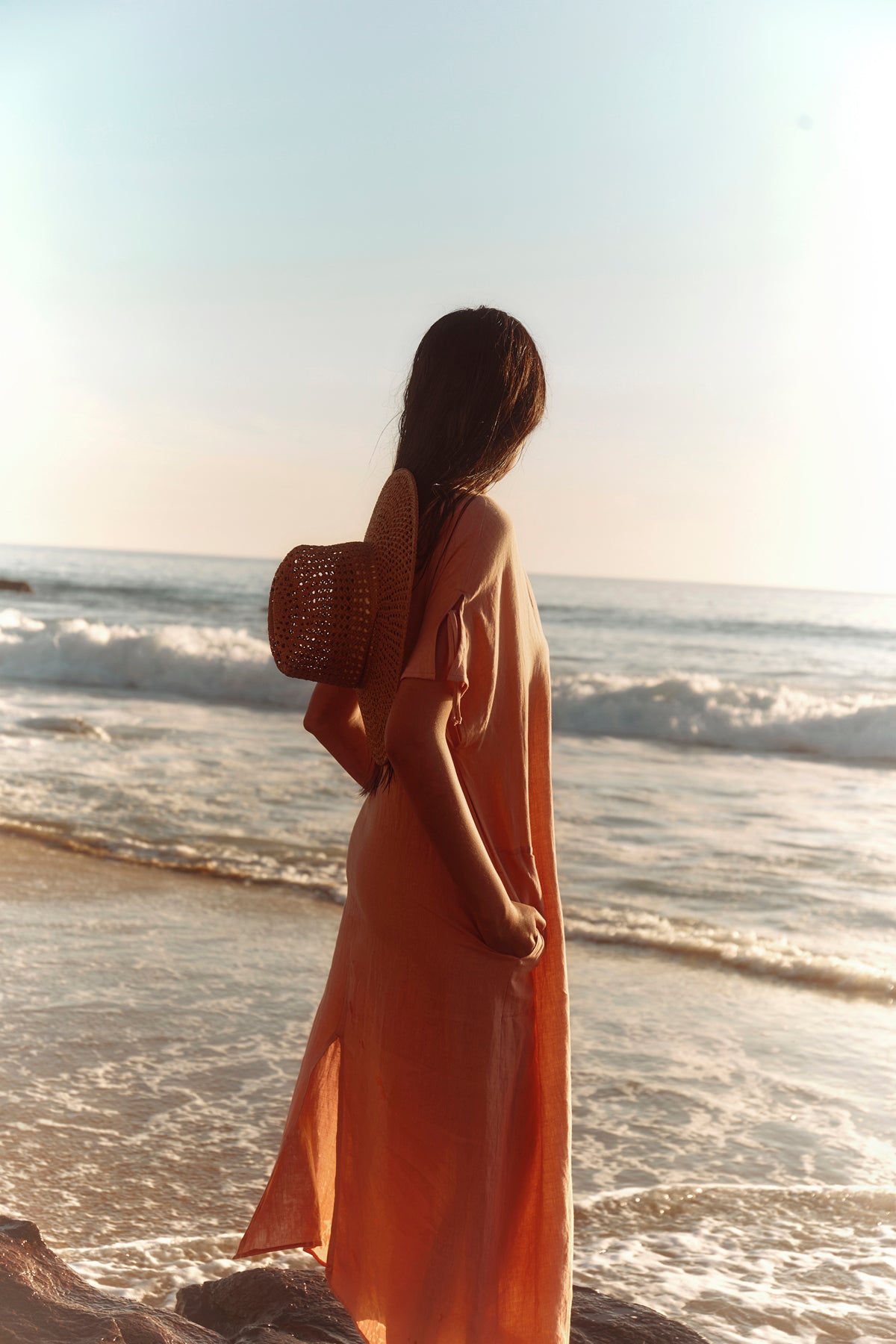 Wearing the MAEVE LINEN DRESS by Velvet by Graham & Spencer, a person stands on the beach gazing at the ocean—a stylish choice for casual seaside outings.