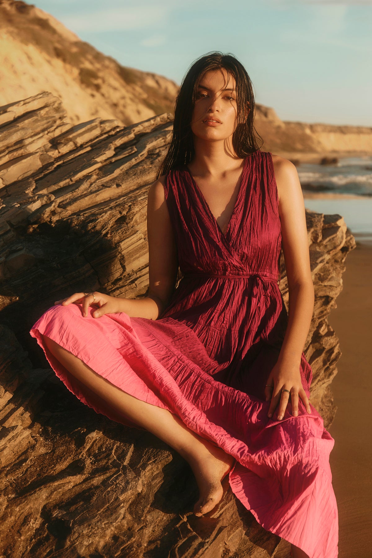 A woman in the MARILYN DRESS by Velvet by Graham & Spencer, a sleeveless dip-dye crinkle voile maxi with a wrap front V-neck, sits on layered rocks at the beach with ocean and cliffs behind her.