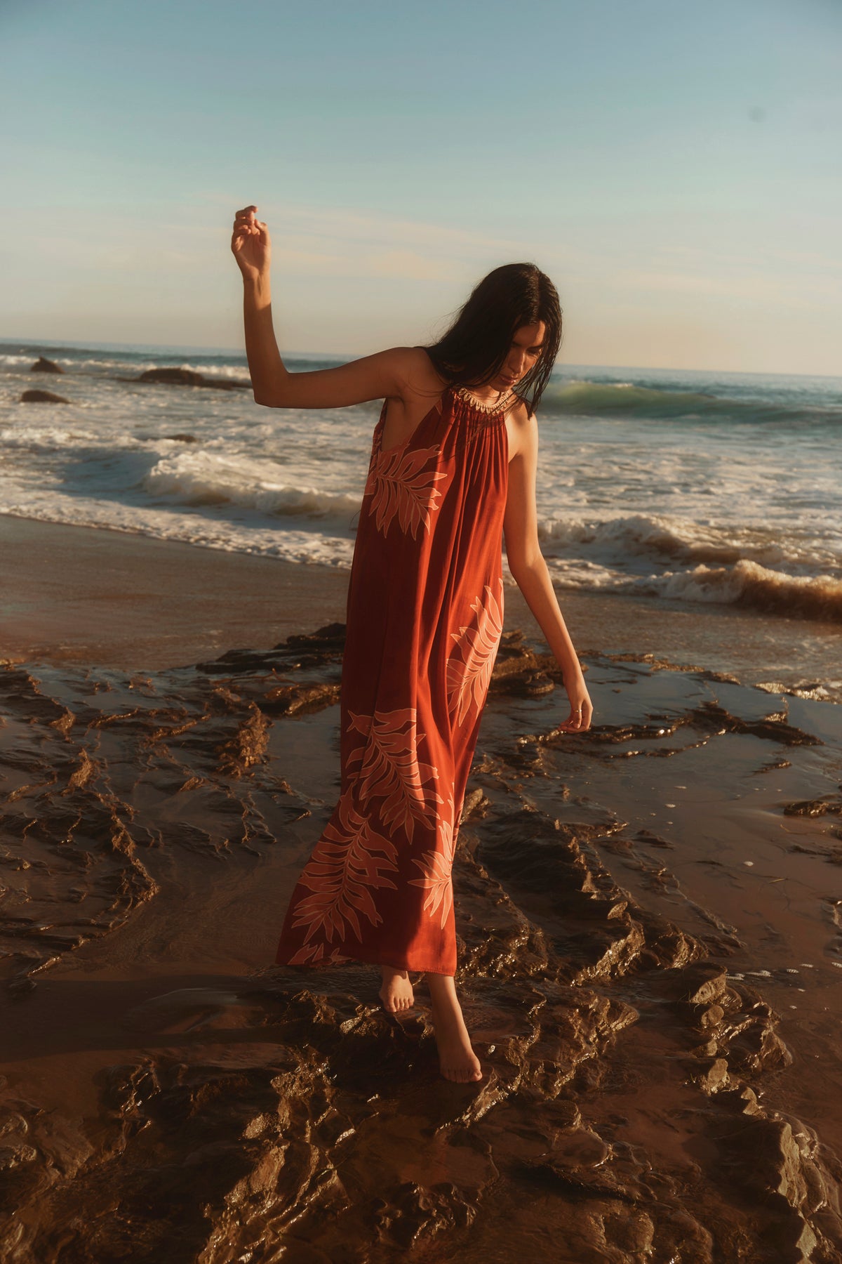 A woman in the ODESSA DRESS by Velvet by Graham & Spencer, a long halter-style red dress with leaf patterns, walks barefoot on a rocky beach near the shoreline at sunset.-39123393511617