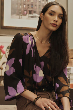 A woman is sitting on a chair in a room, wearing an ISRA PRINTED V-NECK BLOUSE by Velvet by Graham & Spencer with a timeless look and V-neckline.