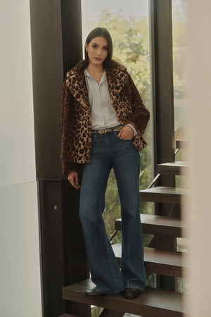 A woman standing on a staircase, wearing the AMANI LEOPARD LUX FAUX FUR JACKET by Velvet by Graham & Spencer, a white blouse, and flared jeans, with sunlight filtering through large windows in the background. The ensemble is a perfect addition to any fall wardrobe.
