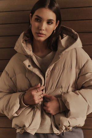 A woman in a Velvet by Graham & Spencer RAYLIN CROPPED PUFFER JACKET leaning against a wooden wall.