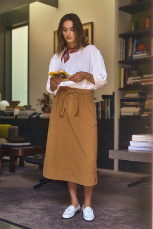 A woman stands in a room holding a book, wearing a white shirt, the versatile KENNEDY CORDUROY SKIRT from Velvet by Graham & Spencer in brown, and white shoes. Bookshelves are visible in the background.