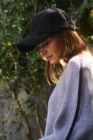 Someone outdoors, surrounded by greenery, wears the Velvet by Graham & Spencer SHERPA CAP—a textured black cold-weather baseball cap—paired with a light gray sweater, looking downward.