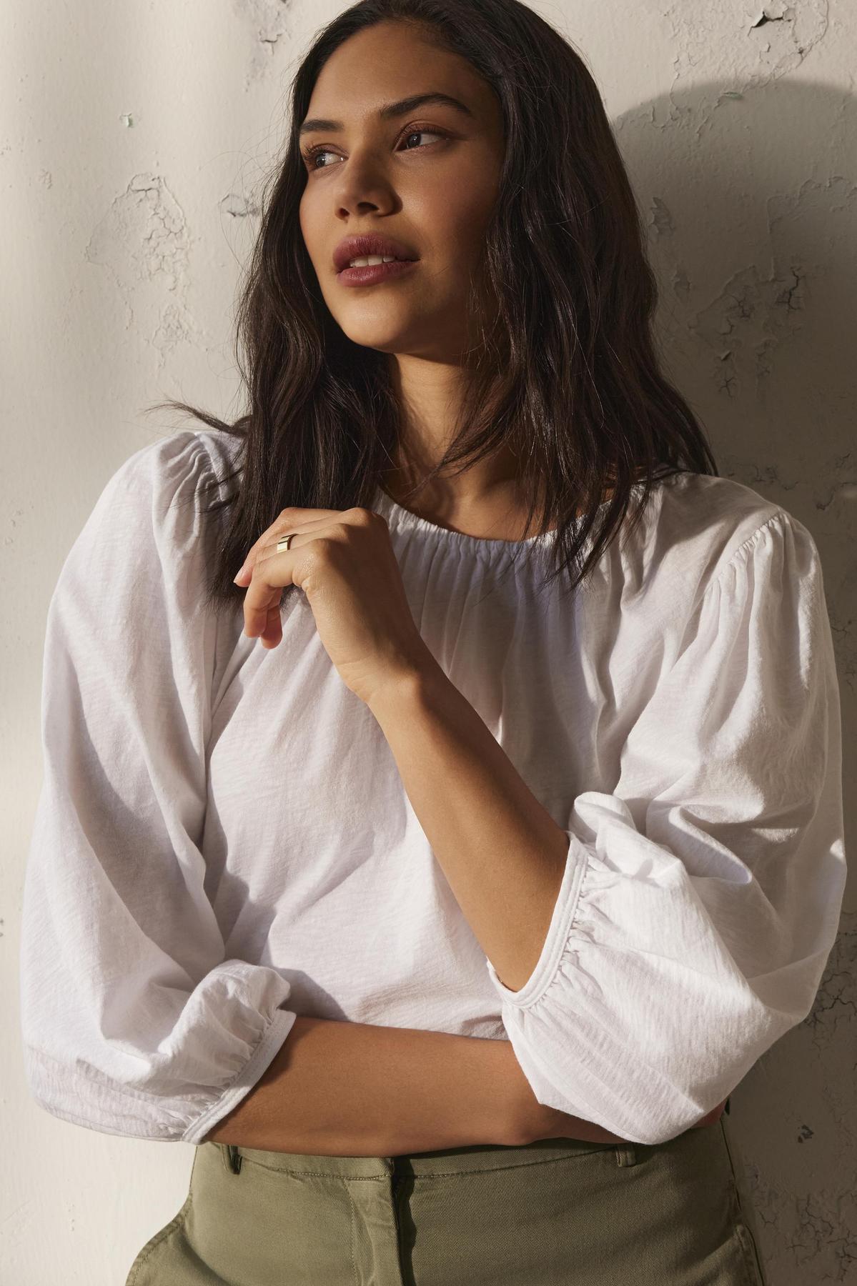 A woman with long brown hair stands against a textured white wall, wearing the CARENA TEE by Velvet by Graham & Spencer and green pants.-38499462906049