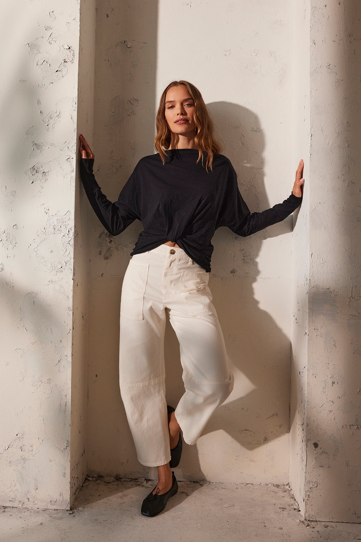 A woman in the HALLIE TEE by Velvet by Graham & Spencer, featuring black long dolman sleeves, pairs it with white pants and chic black flat shoes as she leans against a textured wall. Her hands gently touch the sides.
