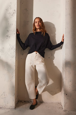 A woman in the HALLIE TEE by Velvet by Graham & Spencer, featuring black long dolman sleeves, pairs it with white pants and chic black flat shoes as she leans against a textured wall. Her hands gently touch the sides.