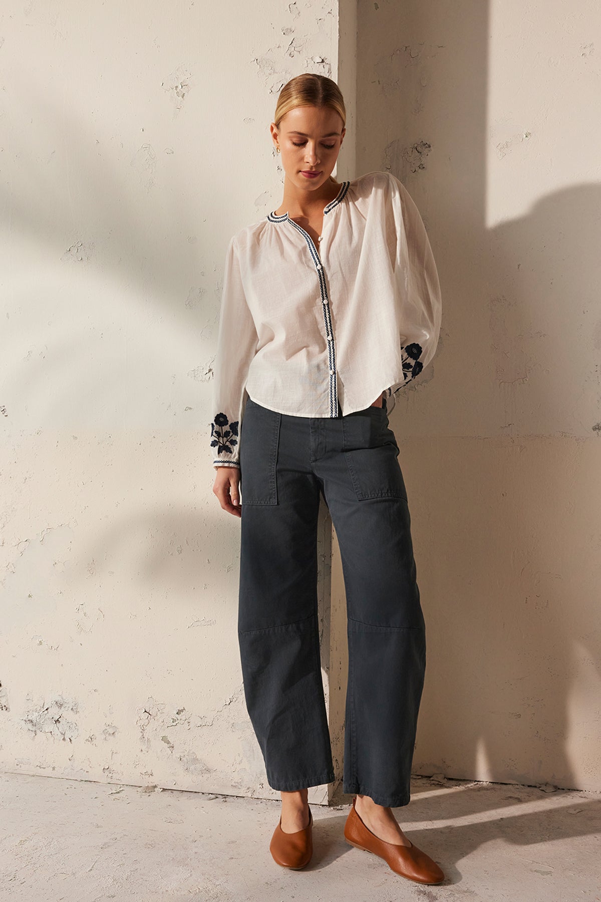 A person wearing the RAHWA TOP by Velvet by Graham & Spencer, featuring floral embroidery on the sleeves, stands against a textured wall in dark cotton pants and brown shoes. They have short hair and their hands are behind their back while looking down.-38866415845569