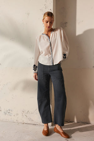A person wearing the RAHWA TOP by Velvet by Graham & Spencer, featuring floral embroidery on the sleeves, stands against a textured wall in dark cotton pants and brown shoes. They have short hair and their hands are behind their back while looking down.